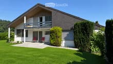 Two-story house with a modern design, featuring a slanted roof, large windows, and a balcony on the upper level. The front yard includes a well-maintained lawn, trimmed hedges, and a small patio area with two red chairs and a table.
