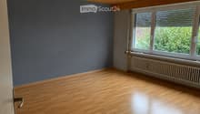 Empty room with wooden flooring, grey walls, large window with blinds, radiator, and a door with a silver handle.