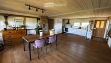 Open-plan living and dining area with wooden floors, exposed wooden beams, a dining table with chairs, a cabinet, and a modern kitchen area with white cabinets and stainless steel appliances.