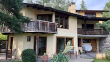 Two-story house with a wooden balcony, large windows, stone chimney, and outdoor seating area with a table and chairs.
