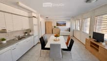 Bright, modern open-plan living and dining area with a fully equipped kitchen, dining table with chairs, TV stand, and a separate bedroom area with a bed and wardrobe.