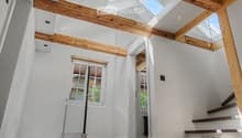 Bright, airy hallway with exposed wooden beams, skylights, and a staircase leading upwards. White walls and wooden flooring.