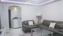 Modern living room with a grey sectional sofa, white throw pillows, a glass coffee table, a floor lamp, and a small white cabinet in the background.