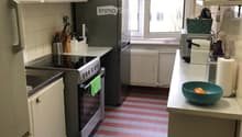 A compact kitchen with white cabinets, stainless steel appliances, and a striped red and white tiled floor. Features include a refrigerator, oven, sink, and various kitchen items on the counter.