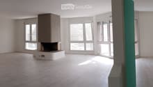 Empty room with wooden flooring, white walls, large windows with blinds, and a fireplace in the center.
