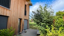 A modern wooden house with a pathway leading to the entrance, surrounded by lush greenery and plants.