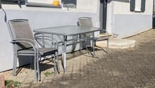 Outdoor seating area with a metal table and four chairs, situated next to a building with shutters and a small awning.