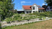 Two-story modern house with red-tiled roof, large windows, and a raised ground floor surrounded by a stone retaining wall and greenery.