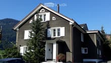 Two-story house with dark brown siding, white shutters, and a raised ground floor entrance. A small garden area with potted plants is visible near the entrance.
