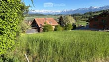 A scenic view of a grassy field with two houses in the background, surrounded by lush greenery and distant snow-capped mountains.