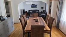 Dining area with a wooden dining table and chairs, a living area with a sofa and armchair, and a television. The room features wooden flooring and white walls.