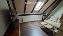 An attic room with wooden floors, exposed wooden beams, and a skylight. It features a sofa, coffee table, a wicker chair, a screen, and a small table with items on it.