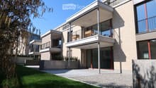 Modern multi-story building with balconies, large windows, and a clean, contemporary design. Features include a raised ground floor, a well-maintained lawn, and a paved pathway leading to the entrance.