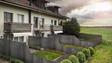 Two-story house with a tiled roof, multiple windows, and balconies. The surrounding area includes a well-maintained garden with a grey wooden fence, shrubs, and flower beds.
