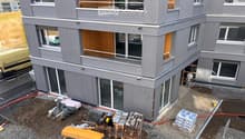 Modern two-story building with grey siding, large windows, and balconies. Construction materials and tools are visible on the ground.