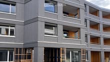 Modern multi-story building with grey wooden exterior, large windows, and balconies. Construction materials and scaffolding are visible in the foreground.