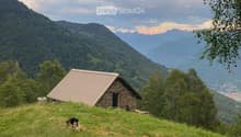 A small stone house with a metal roof sits on a grassy hillside, surrounded by trees and mountains.