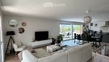 Bright, modern living room with large windows and a balcony. Features a grey sectional sofa, coffee table, armchair, and a dining area with a table and chairs.