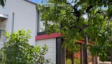 Modern two-story house with white walls, red accents, large windows, and a balcony. Surrounded by lush greenery and a stack of firewood near the entrance.