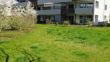 Modern multi-story residential building with balconies, surrounded by a well-maintained lawn and small garden area.