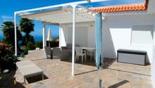 Outdoor patio with stone tiles, white pergola, sofa, table, chairs, and umbrella, overlooking the ocean.