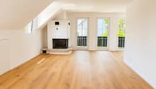 Bright, unfurnished room with wooden flooring, white walls, and a fireplace. Three large windows and doors lead to an outdoor area.