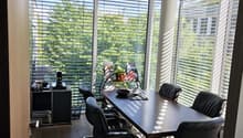 Modern office space with large windows, wooden floor, black leather chairs, a long table, a small cabinet, and a potted plant.
