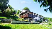 Two-story house with a raised ground floor, white exterior, wooden balcony, and surrounded by greenery.