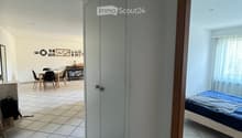 A hallway leads to a furnished room with a bed, a dining area with a table and chairs, and a small cabinet.