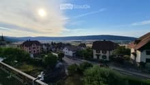 Balcony overlooking a small town with houses, a road, and distant hills under a clear sky.