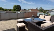 A modern rooftop terrace with a wicker seating set, glass table, and lantern, overlooking a residential area with trees and houses.