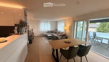 Open-plan living and dining area with light wood flooring, white walls, large windows, and a sliding glass door leading to a balcony. Features a grey sectional sofa, coffee table, dining table with chairs, and a kitchen counter with a vase and plant.