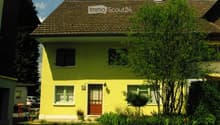 Single-story yellow house with a brown tiled roof, white-framed windows, and a small front garden with trees and shrubs.