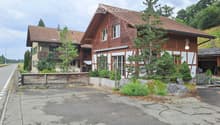Two-story wooden house with red-tiled roof, multiple windows, and a small balcony on the left side. The house is surrounded by greenery and a wooden fence.
