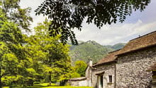 Stone house with a thatched roof, surrounded by lush green trees and a grassy field, with mountains in the background.