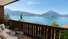 Balcony with wooden railing, overlooking a lake and mountains, furnished with a sofa, small table, candle, and potted plants.
