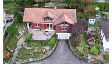 A two-story house with a red-tiled roof and red shutters, surrounded by a well-maintained garden and driveway. The house features a garage and a small patio area with outdoor furniture.