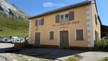 A small, yellow building with brown shutters and a wooden door, labeled "COL du SIMPLON-PASS" with an altitude of 2005 m. It is situated near a parking area with several vehicles and a mountainous backdrop.