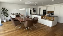Furnished Room. Modern dining area with wooden table, brown chairs, and a white cabinet. A plant and flowers add a touch of nature. The kitchen has a counter, microwave, and oven.