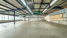 Large, empty industrial-style space with concrete floors, exposed metal beams, and large windows allowing natural light. Minimal furnishings, including a few stacked crates and a small table in the background.