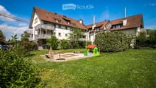 A well-maintained garden area with a small playhouse, benches, and a table, surrounded by a two-story residential building with balconies and a red-tiled roof.