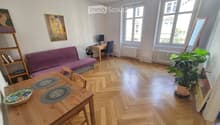 A cozy living room with wooden flooring, a purple sofa, a wooden dining table with placemats and a decorative bowl, a small desk with a laptop, a potted plant, and artwork on the walls.