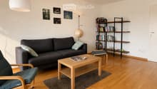 Modern living room with wooden flooring, grey sofa, wooden coffee table, armchair, bookshelf, and decorative picture frames on the wall.
