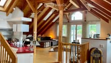 Open-plan attic space with exposed wooden beams, high ceilings, and skylights. Features a living area with a sofa, coffee table, and decorative items, a kitchen area with a red countertop, and a staircase leading to another level.