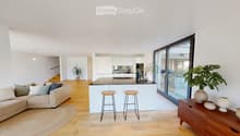 Open-plan living and dining area with a modern kitchen island, sofa, bar stools, and decorative vases. Large windows offer a view of the outdoor space.