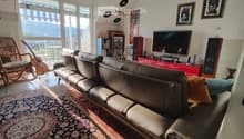 Modern living room with large windows offering a scenic view, featuring a leather sofa, ottoman, wicker chair, guitar, bookshelf, television, speakers, and framed artwork.