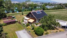 A two-story house with solar panels on the roof, surrounded by a well-maintained garden, a small wooden shed, and a view of a lake and distant mountains.
