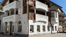 A multi-story building with white walls, wooden accents, and balconies. The building features a sign reading "PIZZERIA" and has multiple windows with curtains. The ground is paved with cobblestones.