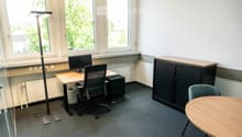 Office space with a desk, chair, computer monitor, cabinet, round table, and floor lamp. Large windows provide natural light.