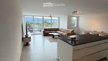 Modern open-plan living and dining area with a sectional sofa, coffee table, kitchen island, and large windows offering a scenic view.